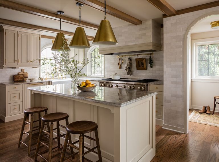 At the stove alcove, a Wolf range sits beneath a tiled hood with a Vent-A-Hood insert. The sink is from Shaws and the faucet from Rohl. The tile throughout is the Cloe Tile in White by Bedrosians. Visual Comfort pendants hang over the new island, which is a family hub of cooking and conversation, with Sea Pearl quartzite counters and vintage stools.