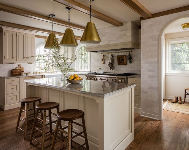 At the stove alcove, a Wolf range sits beneath a tiled hood with a Vent-A-Hood insert. The sink is from Shaws and the faucet from Rohl. The tile throughout is the Cloe Tile in White by Bedrosians. Visual Comfort pendants hang over the new island, which is a family hub of cooking and conversation, with Sea Pearl quartzite counters and vintage stools.