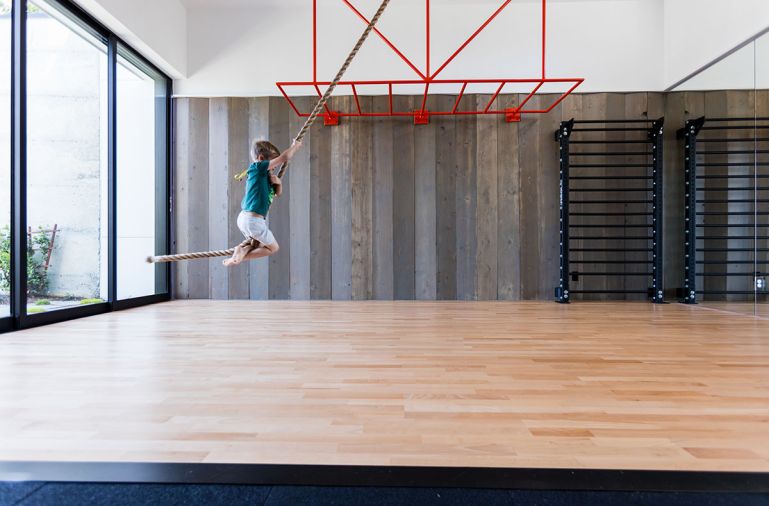 The gym’s hardwood flooring is from Fitness Flooring. The fitness space comes equipped with bright orange monkey bars, a climbing rope, and a water fountain. One side of the gym is covered in the original exterior wood siding reclaimed from the prior home.