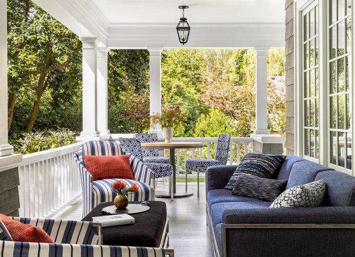 A 1920s photo revealed the current property sits on two lots, with an adjacent home apparently razed and lot purchased so this porch could be added to the south side. During trip to San Francisco, homeowners sat in the Sutherland showroom and chose the Perennial indoor-outdoor fabrics for each. Pops of blues, blacks, and orange pair with black indoor-outdoor mohair Ottoman. Orange pillows from Susan Mills at the Seattle Design Center.