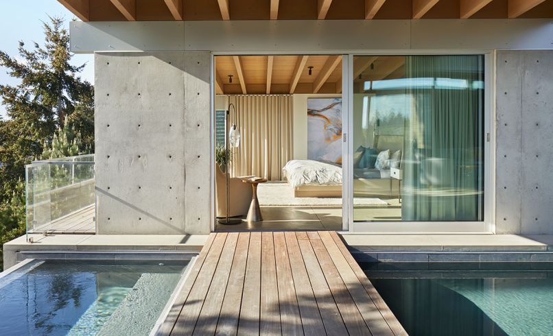 An exterior Ipe footbridge leads to the primary suite, which can be reached via an interior hallway. The footbridge separates the lap pool on the right from the spa on the left. In the bedroom, custom bed frame and window coverings are from Garret Cord Werner Architects and Interior Designers.