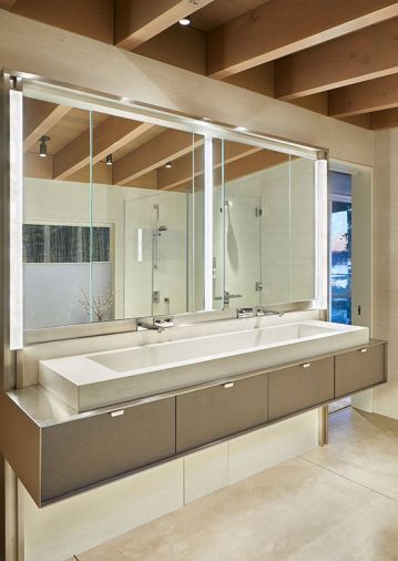 In the primary bathroom, Mercer Builders made a custom mirror cabinet on-site with opaque glass light from Stephen Hirt and glass from Distinctive Glass. The custom floating vanity features a stone basin sink atop a stainless-steel bar counter.