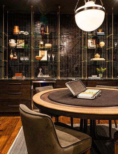 The downstairs whiskey bar has custom shelving by Forsyth Metal Works joined with black wall tile, a game table by Burke Decor, and chairs from Four Hands.