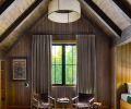 The lodge-style bunk room under a soaring gabled roof is wrapped in dark-stained wood, with window coverings from Cindy’s Window Fashions. Ottomans from Grandin Road and an RH rug offer a pop of pattern, and the ceiling light is the Dunne Wide Pendant from Visual Comfort.