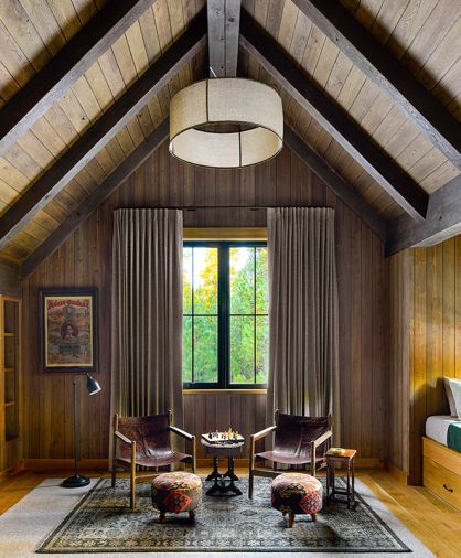 The lodge-style bunk room under a soaring gabled roof is wrapped in dark-stained wood, with window coverings from Cindy’s Window Fashions. Ottomans from Grandin Road and an RH rug offer a pop of pattern, and the ceiling light is the Dunne Wide Pendant from Visual Comfort.