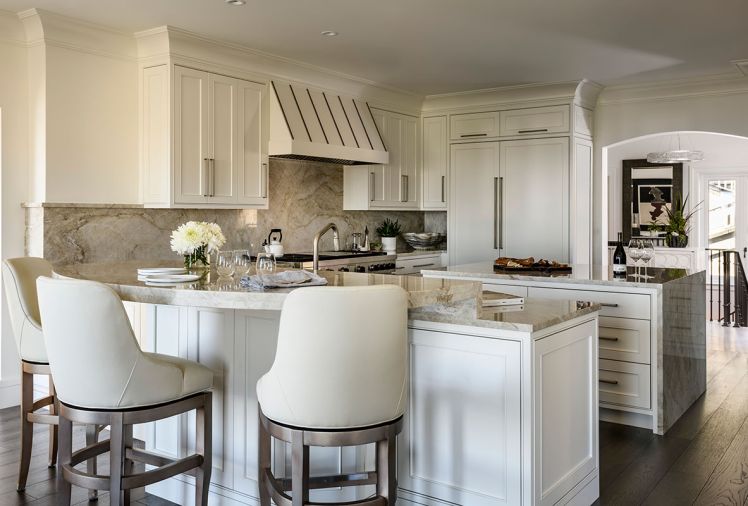 In the kitchen, custom, paint-grade cabinetry from Interior Environments is covered in “White Dove” color. A Taj Mahal polished countertop and backsplash is from Marmo e Granito. At the bar, find Lauren Memory Swivel Barstools by Jessica Charles. All appliances are Wolf Sub-Zero.