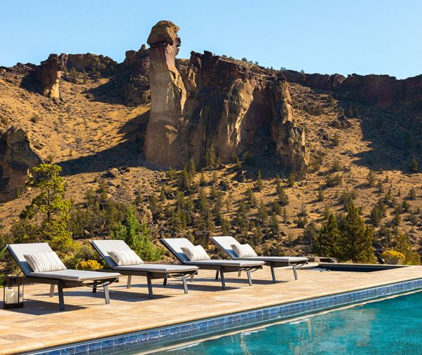 A row of Santa Barbara Outdoor Metal Chaises from Williams Sonoma Home sit atop the travertine pool surround. Just behind is the iconic Monkey Face in Smith Rock State Park.
