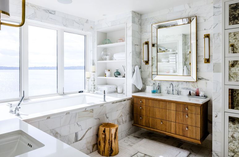 Large Kohler Underscore Rectangle tub is faced with Walker Zanger Studio Moderne™ Calacatta honed from United Tile. Keely Tall Pivoting sconces trimmed in hand-rubbed antique brass are grounded by warm Winterwood Custom vanity. Organic White counter from Caesarstone.