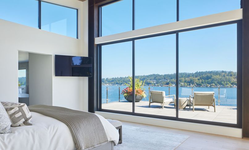 Natural light floods the primary suite through expansive Marvin windows from Cherry Creek Windows & Doors. Custom deck railings by AGS Stainless Inc. offer clear water views.