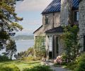 The reclaimed granite quarry tailings walkway meanders toward a grand limestone fireplace located near the entrance, all crowned by a steeply pitched metal roof. A cozy partially enclosed patio is oriented toward a view of the peninsula beyond.