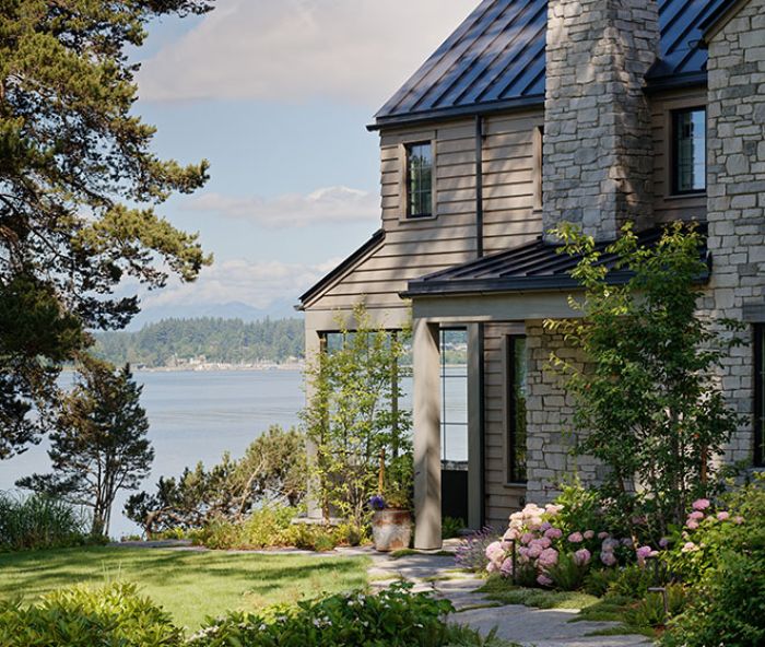 The reclaimed granite quarry tailings walkway meanders toward a grand limestone fireplace located near the entrance, all crowned by a steeply pitched metal roof. A cozy partially enclosed patio is oriented toward a view of the peninsula beyond.