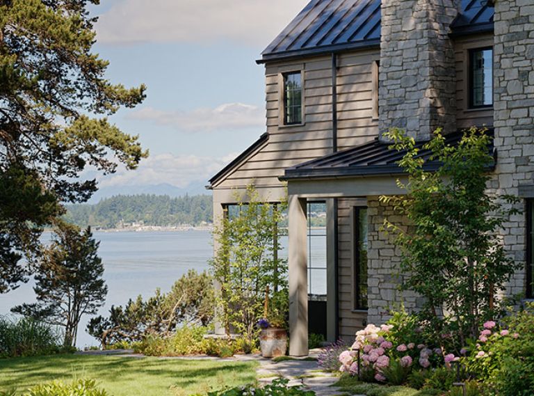 The reclaimed granite quarry tailings walkway meanders toward a grand limestone fireplace located near the entrance, all crowned by a steeply pitched metal roof. A cozy partially enclosed patio is oriented toward a view of the peninsula beyond.