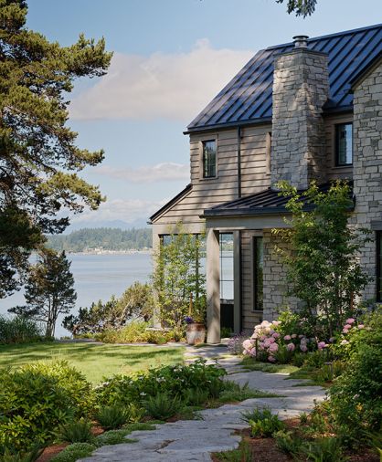 The reclaimed granite quarry tailings walkway meanders toward a grand limestone fireplace located near the entrance, all crowned by a steeply pitched metal roof. A cozy partially enclosed patio is oriented toward a view of the peninsula beyond.