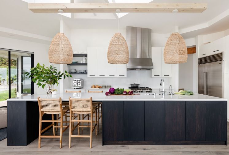 An oak island with custom ebony dye and quartz countertop pops against Benjamin Moore’s “Simply White” walls. Custom cabinetry throughout by Juggernaut Woodworking. Serena & Lily rattan pendants suspended below the wagon-wheel skylight echo teak and rattan stools below.