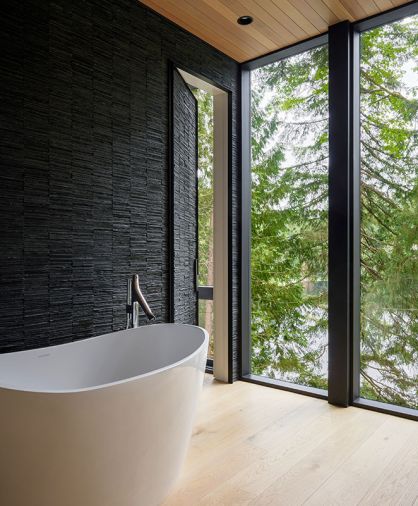 An Amalfi Slipper Tub in gloss white and a Hansgrohe Axor Stark Organic Tub Filler sit beside Porcelanosa Skyline Linear Broken Dark Slate and forest views, blending calm and artistry.