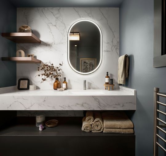 The wellness studio bathroom has a Dekton Entzo counter with full-height backsplash, a Kichler Menillo LED Mirror, a radiant heat towel bar and custom wood shelving.