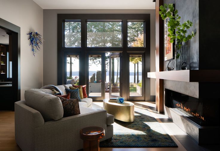 The corner fireplace insert is the Ortal 77 RC gas fireplace from Custom Fire Art, with a Taylor Made Custom Cuddle Sectional in front. A Caracole “Come Oval Here” coffee table reflects natural light via a sleek metallic sheen.