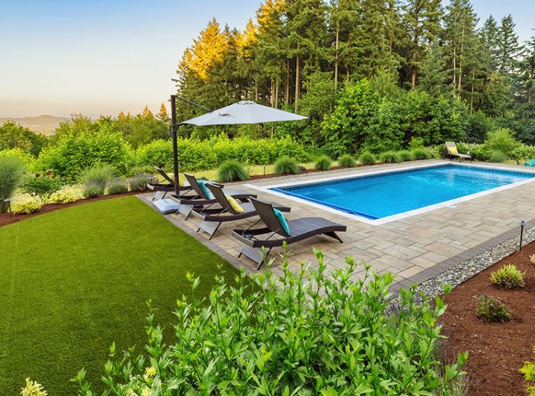 Hameln Fountain Grass softens the length of the pool trimmed in Western Interlock pavers and a river rock border. An Azurri Blue Satin Hibiscus flourishes in the foreground.