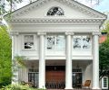 A classic fan light window crowns four massive Ionic columns restored by Arciform. The brick chimney is flanked by two pairs of restored French doors on the side porch.
