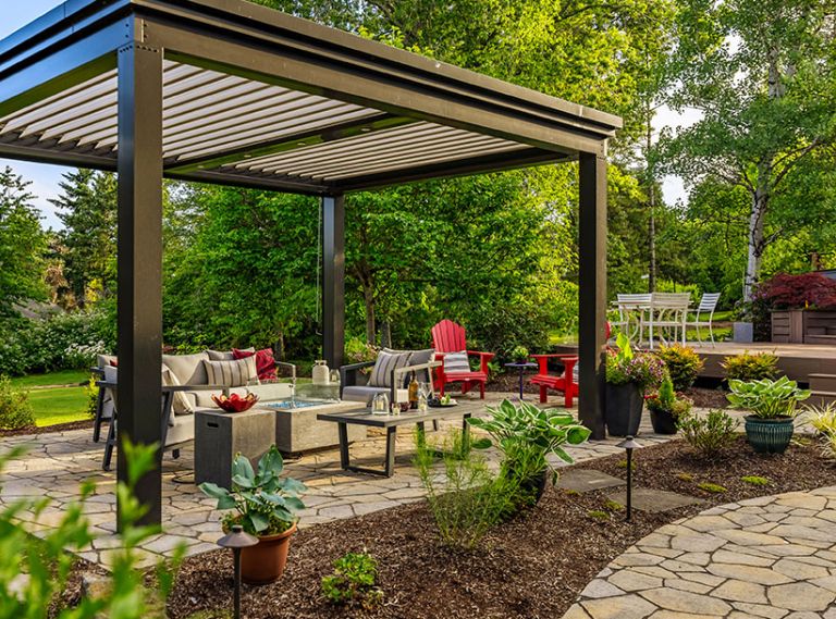 The freestanding louvered pergola in &ldquo;Onyx&rdquo; from The Luxury Pergola adds year-round protection to seating area and firepit which sits on top of Panorama Flag pavers. The lighting along the pathway adds dimension at night.