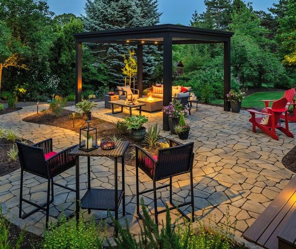 Thoughtfully placed uplights give the surrounding trees a soft glow as night falls. The homeowner&rsquo;s herb garden includes rosemary and oregano, and nearby plantings include Snowball Viburnum, Hostas, and Liriope Lilyturf.