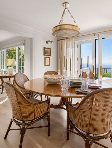 Soothing wood-based furnishings are crowned by an Urban Electric chandelier. The Johnathan Charles table is surrounded by wicker chairs and French doors dressed with drapery open onto an expanded deck.