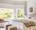 Peaceful furnishings for the bedroom include a vintage Turkish rug, textured stools, Roman shades, and an inviting bedframe. &ldquo;Pale Oak&rdquo; by Benjamin Moore dresses the walls and a reflective Rejuvenation light fixture shines overhead.