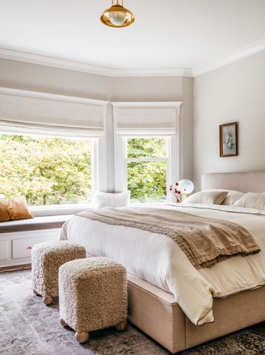 Peaceful furnishings for the bedroom include a vintage Turkish rug, textured stools, Roman shades, and an inviting bedframe. &ldquo;Pale Oak&rdquo; by Benjamin Moore dresses the walls and a reflective Rejuvenation light fixture shines overhead.
