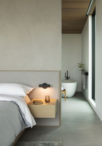ABOVE The primary bedroom includes a custom bed with headboard in &ldquo;Nitty Witty&rdquo; fabric from Perennial. Lit floating bedside tables add to the calming ambiance. A view into the bathroom includes the Essentia, Bain Ultra soaking tub.