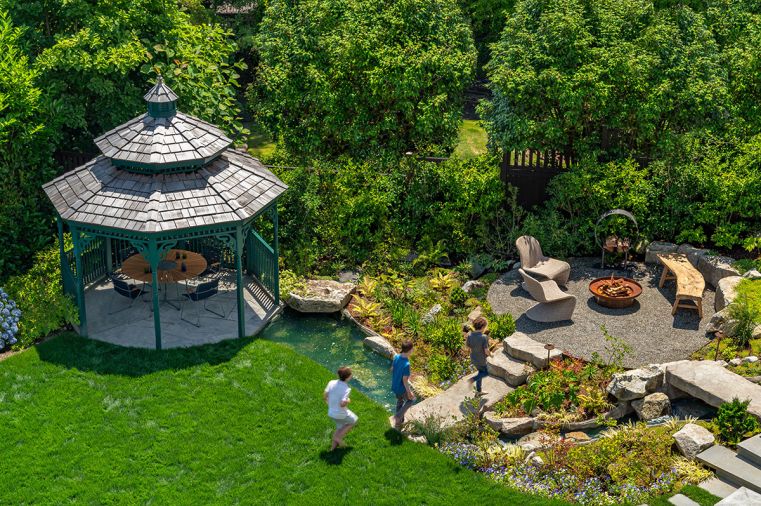 In the backyard, a stream runs to the ten-foot octagon wood gazebo from Amish Country Gazebos. The foot bridges over the stream were custom-painted to look like stone. In the fire pit area, the firebowl with optional tabletop is from Paloform.