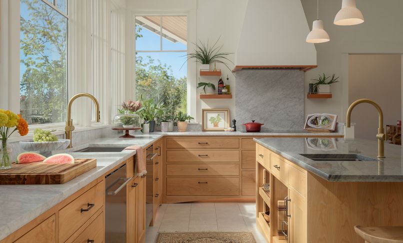 The light and airy kitchen gives a cheerful vibe even on overcast days, with a tactile palette of alder cabinetry, Carrara marble, and Kohler brass fixtures from Ferguson Home. Maple hardwood flooring in the adjoining space reiterates the wood tones in the kitchen.
