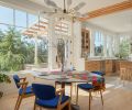 Windows from Island Sash & Door create a year-round connection to the outdoors, while the dining area pairs a pedestal table with vibrant blue chairs and a Kanova Lighting chandelier from Globe Lighting.