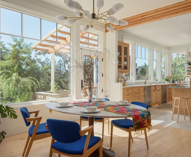 Windows from Island Sash & Door create a year-round connection to the outdoors, while the dining area pairs a pedestal table with vibrant blue chairs and a Kanova Lighting chandelier from Globe Lighting.