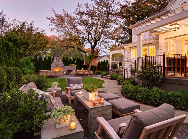 Pergola covered decks are a transition between inside and out in this Portland home. At ground level, a winding bluestone path connects the house to the outdoor rooms. The homeowners wanted the space to fulfill three roles: provide one area in which to entertain guests or gather with family, a second area with grassy lawn for sports practice and a third more intimate spot for reading or enjoying a quiet conversation. Furniture at the north end was custom designed for the space, while the Gloster loveseat, chairs and stools (pictured left) are from Fishels in Portland.