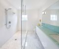 The bathroom offers a mix of textures in the white palette also seen in the kitchen. The wall-hung cabinetry was treated to the same automotive finish as cabinets in the kitchen. White Carrera marble and white pebble tiles cover the floor. On the shower walls, oversized matte white subway tile is stacked linear. The white tones, glass shower wall and a huge mirror make the most of the natural light that enters the bathroom from a small window.