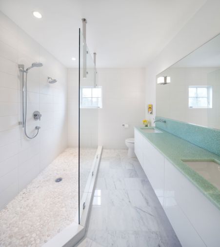 The bathroom offers a mix of textures in the white palette also seen in the kitchen. The wall-hung cabinetry was treated to the same automotive finish as cabinets in the kitchen. White Carrera marble and white pebble tiles cover the floor. On the shower walls, oversized matte white subway tile is stacked linear. The white tones, glass shower wall and a huge mirror make the most of the natural light that enters the bathroom from a small window.