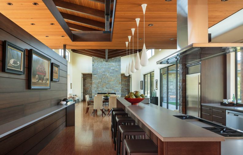 Honed grey quartz counters make cleanup easy in the long galley kitchen. A full height handle stainless Sub-Zero refrigerator is camouflaged by stone on one side and walnut on the other. Crate & Barrel Folio chocolate leather barstools tuck beneath the kitchen counter. Five Goccia H Pendant Lights by Andrea Branzi of Rotaliana from YLighting span the bar adding sparkle to the area also lit from clerestory windows overhead. A dark walnut espresso stain turns glue lams into striking beams. Slotted openings trimmed in walnut stained wood that echoes the screen wall open onto the high ceilings. The kitchen is free of upper cabinets, which are found in the adjacent butler’s pantry and secondary pantry reserved for supplies, tucked behind sliding doors. Floating counters waterfall down to the floor, which feature 12” x 24” cork tiles from Expanko. “We had to go with glued tiles because the runs were too long for plank flooring unless we added thresholds,” says the owner. Dropped ceiling is v-groove Western Red Cedar.