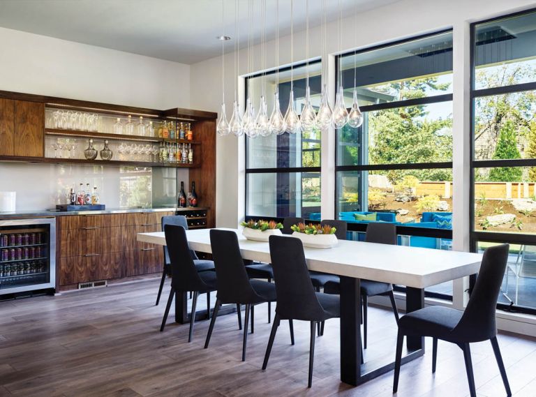The dining area is found in the open communal area, with views out to the spacious backyard, which includes a large patio. Walnut bar cabinetry ties to the adjacent kitchen and includes a moon shadow (gray back painted) backsplash relating to the custom table with concrete skim coat, beverage cooler, wine rack and open glass shelving. Glass pendant chandelier features LED glass bulbs that do not block the view.
