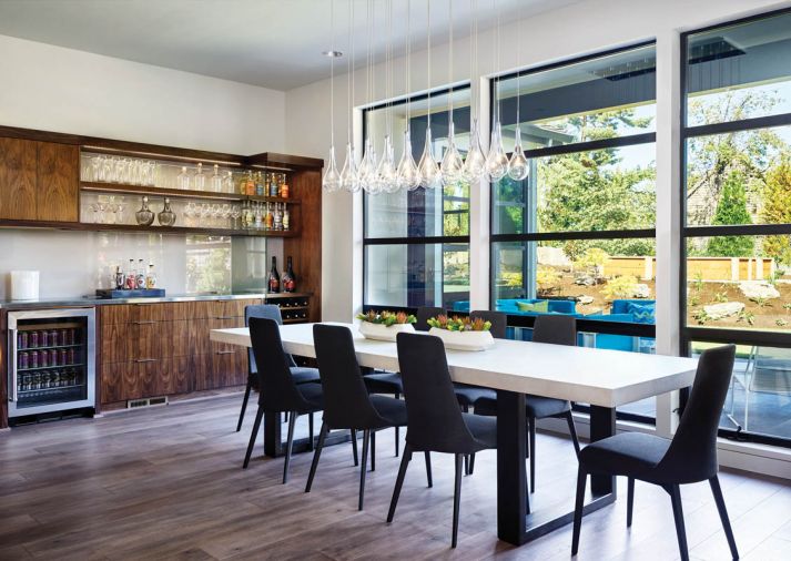 The dining area is found in the open communal area, with views out to the spacious backyard, which includes a large patio. Walnut bar cabinetry ties to the adjacent kitchen and includes a moon shadow (gray back painted) backsplash relating to the custom table with concrete skim coat, beverage cooler, wine rack and open glass shelving. Glass pendant chandelier features LED glass bulbs that do not block the view.