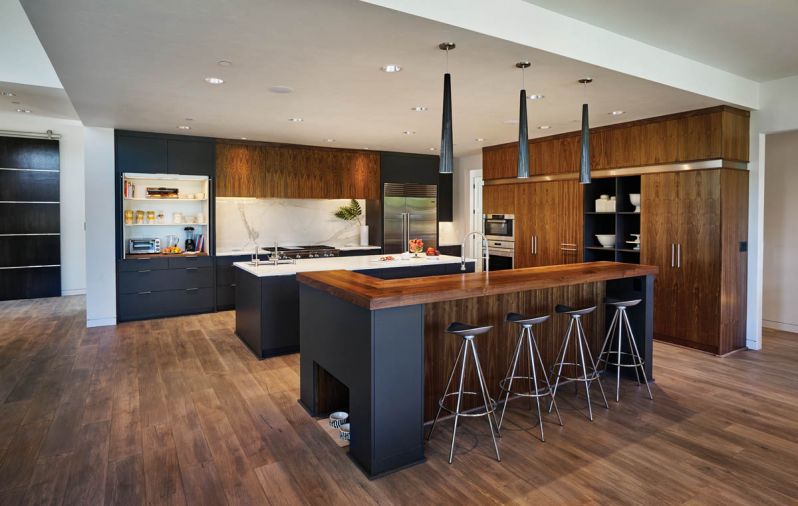 To keep the design contiguous with good visual flow, the use of dark walnut cabinetry along the storage wall was trimmed with a band of stainless steel that reiterates the horizontal stainless steel straps seen on the 10’ barn door with Krownlab hardware leading into the study at left. A breakfast area with appliances to make everything from smoothies to toasted bagels disappears behind receding doors for the rest of the day.