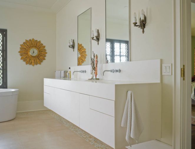 The bathroom lavatory was also custom-designed by Janice Viekman, using solid surface construction that mimics Corian cabinetry. Vola plumbing fixtures were sourced from Best Plumbing, and the wall sconces came from Century Lighting. The overall effect in the bathroom is simultaneously soothing yet visually engaging thanks to the custom-cut shutter and its reflection in the mirrored antique wardrobe.
