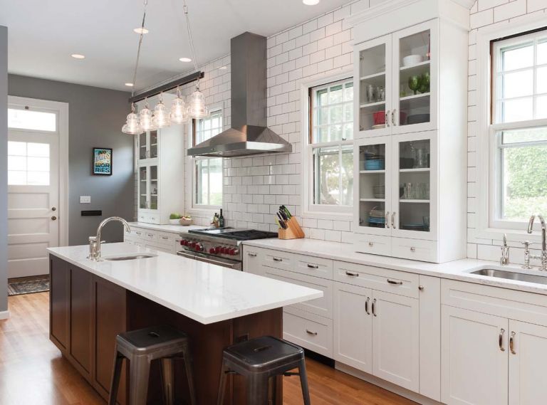 Rohl sink faucets in polished nickel reflect a classic, traditionalist aesthetic that matches the cabinet hardware. Stainless steel appliances echo the same tones, including a professional Wolf range and built-in Sub-Zero refrigerator. A lighting fixture from Troy Lighting with thick, industrial glass shades under-scores the service kitchen aesthetic.