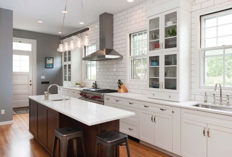 Rohl sink faucets in polished nickel reflect a classic, traditionalist aesthetic that matches the cabinet hardware. Stainless steel appliances echo the same tones, including a professional Wolf range and built-in Sub-Zero refrigerator. A lighting fixture from Troy Lighting with thick, industrial glass shades under-scores the service kitchen aesthetic.